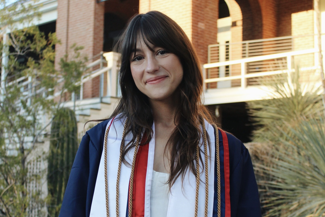 Woman with dark brown hair stands outside a brick building with graduation gown and cords