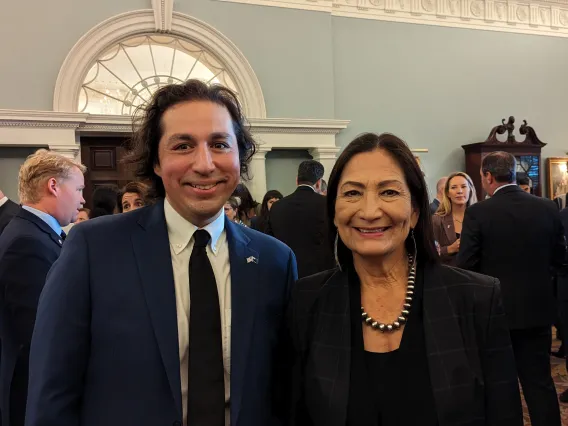 A man in a black suit stands with a woman in a black dress in a large room full of people