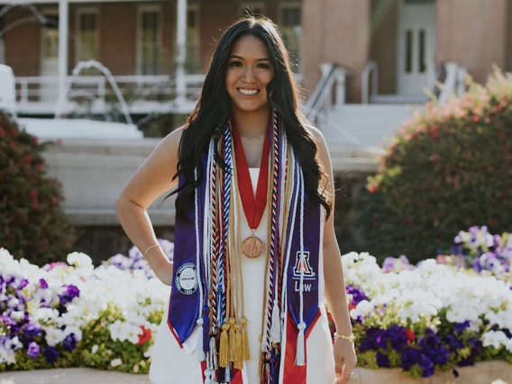 Student with long brown hair stands in front of Old Main, wearing graduation regalia
