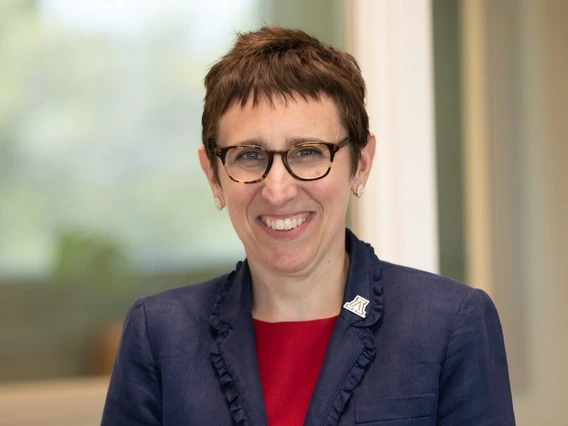 Woman with short brown hair and glasses, wearing a red shirt and blue jacket, sits smiling