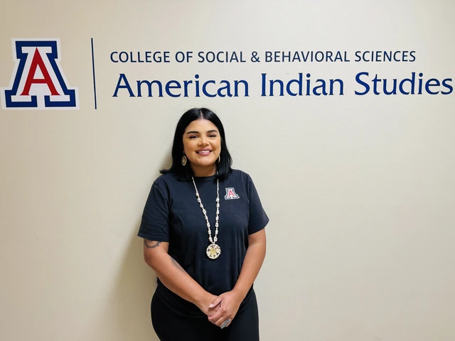 A student with medium length black ahir, wearing a navy blue U of A shirt and Indigenous necklace and ring and standing in front of a sign that says College of Social and Behavioral Sciences American Indian Studies  