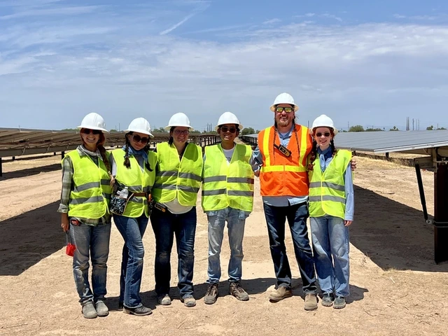 Six people stands outside in front of solar panels and wearing hardhats and safety vests