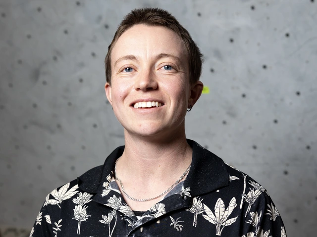 A student with short hair smiles at the camera in front of a light gray rock-climbing wall with bolt holes. They’re wearing a black shirt with a white botanical pattern and a thin chain necklace