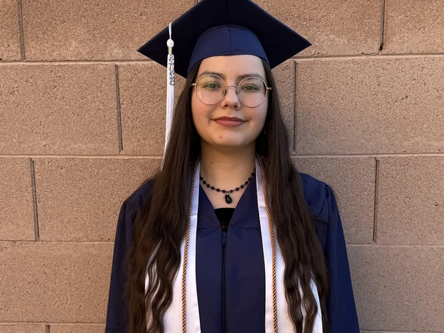 A person wearing a navy blue graduation cap and gown stands in front of a tan brick wall. They have long, wavy dark hair, round glasses, and are wearing a white honor stole and a black beaded necklace. They are looking at the camera with a calm, neutral expression