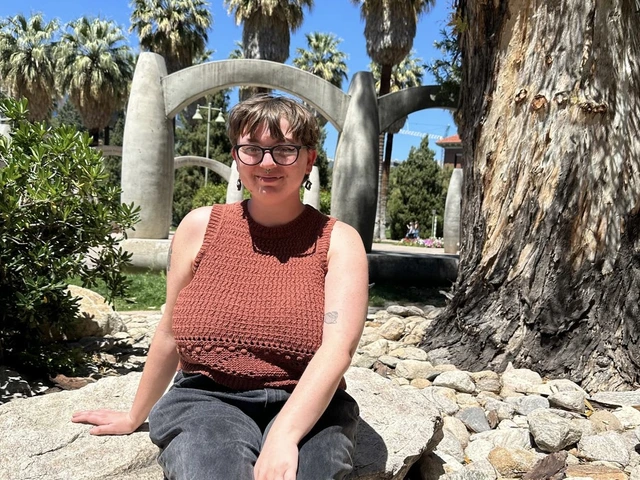 A person with short hair and glasses sits on a rock in a sunny outdoor area. They are wearing a sleeveless rust-colored knit top and dark pants, smiling slightly at the camera. Behind them are large concrete arch sculptures, palm trees, and other greenery