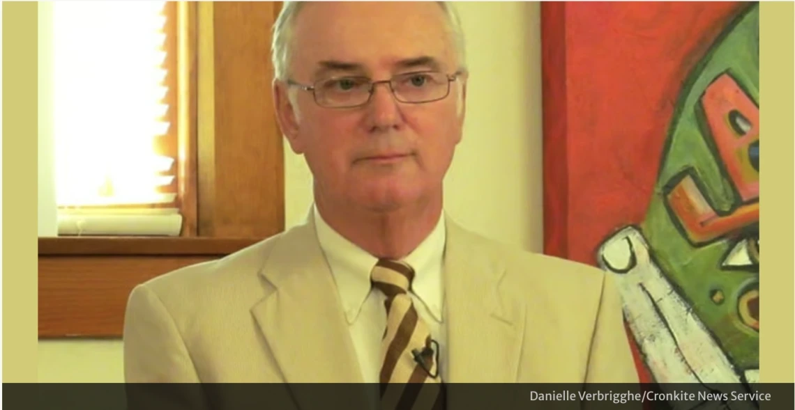 George Cunningham, in a cream colored suit and wearing glasses, stands in front of a brightly colored piece of art