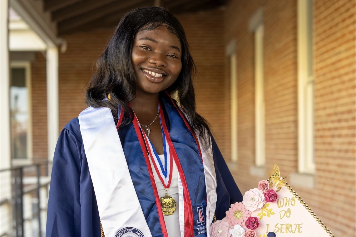 Student stands outside in graduation regalia, holding a graduation cap