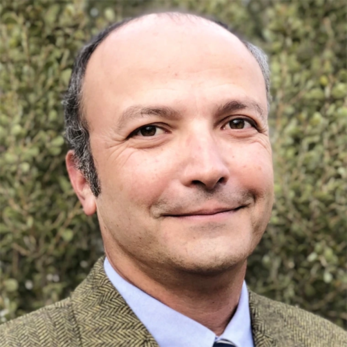 Man in a blue shirt and brown sports coat stand against a hedge, smiling