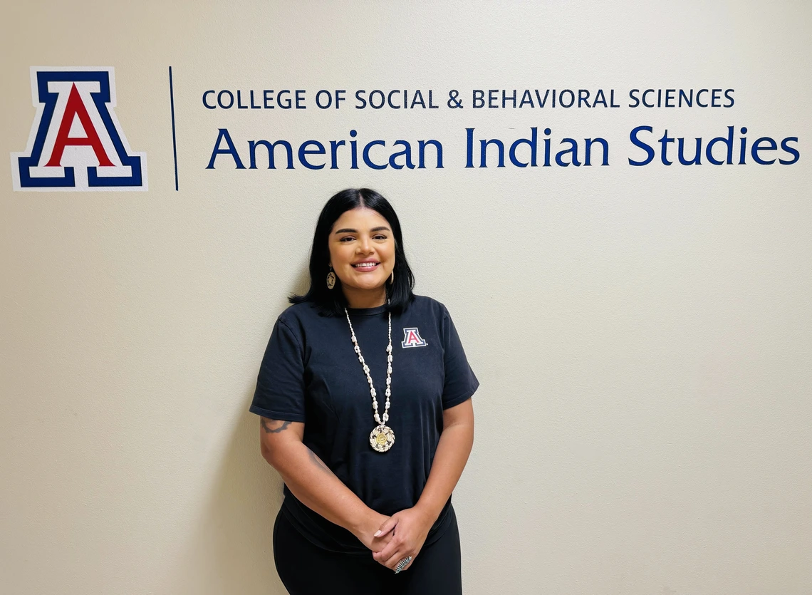 A student with medium length black ahir, wearing a navy blue U of A shirt and Indigenous necklace and ring and standing in front of a sign that says College of Social and Behavioral Sciences American Indian Studies  