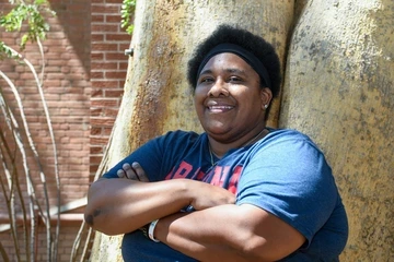A woman wearing a blue and red Arizona tshirt stands smiling with arms crossed against a tree