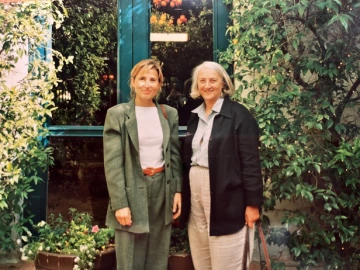 Two women stand next to each other, next to a hedge and in front of a window