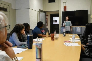 student speaks at the end of a large conference table as students and faculty sit at the table and watch intently