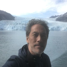Man with a beard taking a selfie with a fjord-like setting in the background