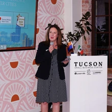 Woman wearing black dress and jacket stands in front of podium and speaks into a microphone