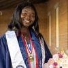 Student stands outside in graduation regalia, holding a graduation cap 