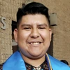 Student stands outside a brick building, wearing graduation regalia
