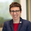 Woman with short brown hair and glasses, wearing a red shirt and blue jacket, sits smiling