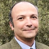 Man in a blue shirt and brown sports coat stand against a hedge, smiling