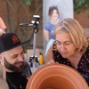 A woman with blonde hair and glasses bends over a piece of pottery to look closely and a man with a black hat and a beard sits near her, looking at the same piece