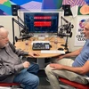 A man in a gray shirt and jeans, wearing headphones, sits across from another man in blue shirt and khakis in a podcast recording studio