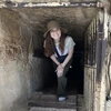 A woman with long brown hair and wearing a hat crouches down, emerging from a mining shaft