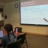 Man with long hair and glasses stands in front of a classroom and projection screen, teaching