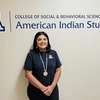 A student with medium length black ahir, wearing a navy blue U of A shirt and Indigenous necklace and ring and standing in front of a sign that says College of Social and Behavioral Sciences American Indian Studies  