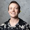 A student with short hair smiles at the camera in front of a light gray rock-climbing wall with bolt holes. They’re wearing a black shirt with a white botanical pattern and a thin chain necklace