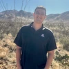 Student with short borwn hair and glasses stands outside in a desert setting and wearing a blue University of Arizona logo polo shirt