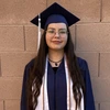 A person wearing a navy blue graduation cap and gown stands in front of a tan brick wall. They have long, wavy dark hair, round glasses, and are wearing a white honor stole and a black beaded necklace. They are looking at the camera with a calm, neutral expression