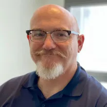 A man with glasses, a mustache and wearing a blue shirt smiles warmly