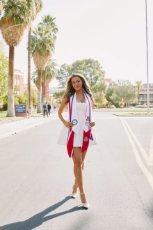 A student wearing white a graduation regalia stands outside with palm trees in the background