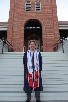 Young man in graduation regalia stands on the steps of Old Main