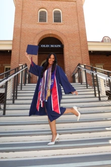 Woman in blue graduation regalia stands on the steps of Old Main and poses with her leg up