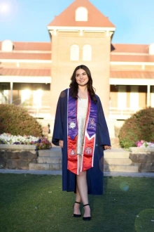 Woman in blue graduation gown stands outside in front of Old Main