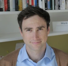 man with brown hair and wearing a blue shirt and brown sweater