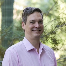 Man in a pink shirt smiles, standing outside