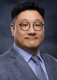 Man with black hair, glasses and a monochromatic suit and shirt 