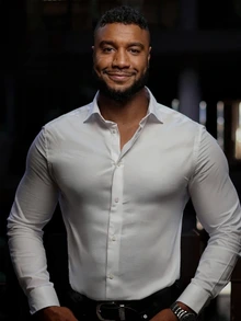 A man with short curly hair and a trimmed beard stands smiling with his hands resting at his sides. He is wearing a fitted white button-down shirt and is photographed indoors against a dim, softly blurred background