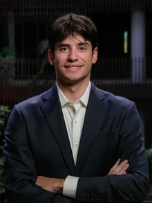 A young man with short dark hair wearing a dark suit jacket and white button-down shirt stands with his arms crossed, smiling slightly. He is photographed indoors against a softly lit, blurred background