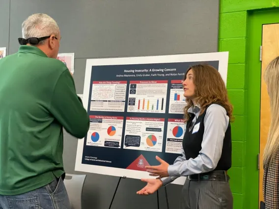 A person with long hair talks to another person who stands in front of a display presentation board