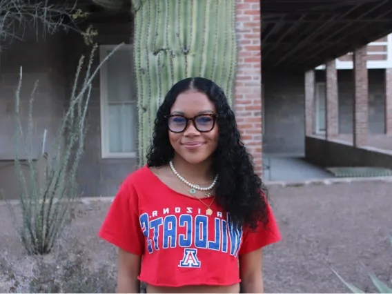 Woman with long dark hair and red shirt standing in front of brick building and a cactus