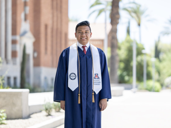 Man with dark hair and blue graduation gown stands in front of a building and palm trees