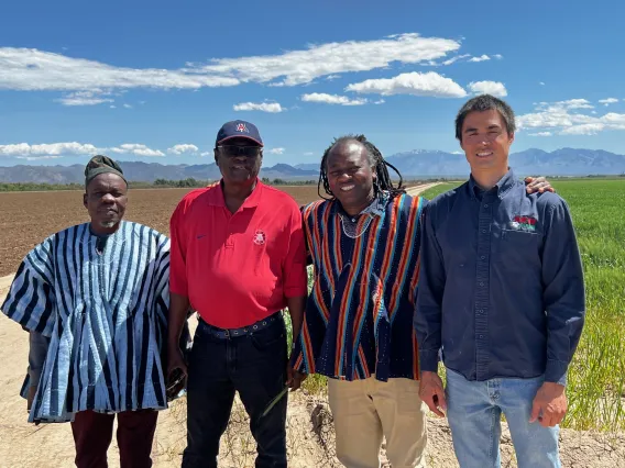 Four men stand outside in the sun with a field and mountains as the backdrop