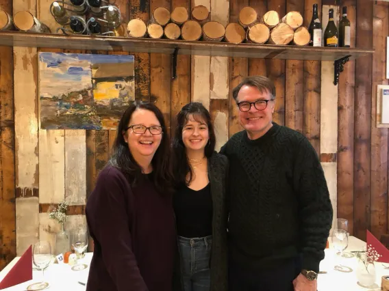 Aurelie Sheehan stands in front of a rustic wood wall with her daughter in the middle and husband to the far right