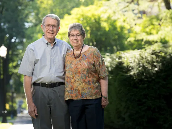 A man and woman stand together outside with sunlit trees in the background