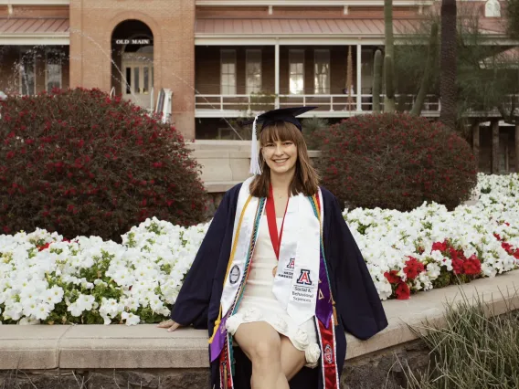 Woman in graduation dress and sandals sits on a brick wall with flowers behind her