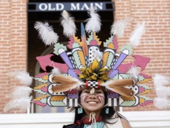 A woman in Indigenous dress stands smiling in front of Old Main building at University of Arizona