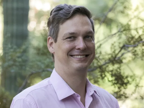 Man with short hair and wearing purple shirt stands outside in the trees