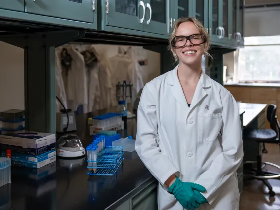 A student with blonde hair, wearing lab glasses and a white lab coat, stands smiling in a lab